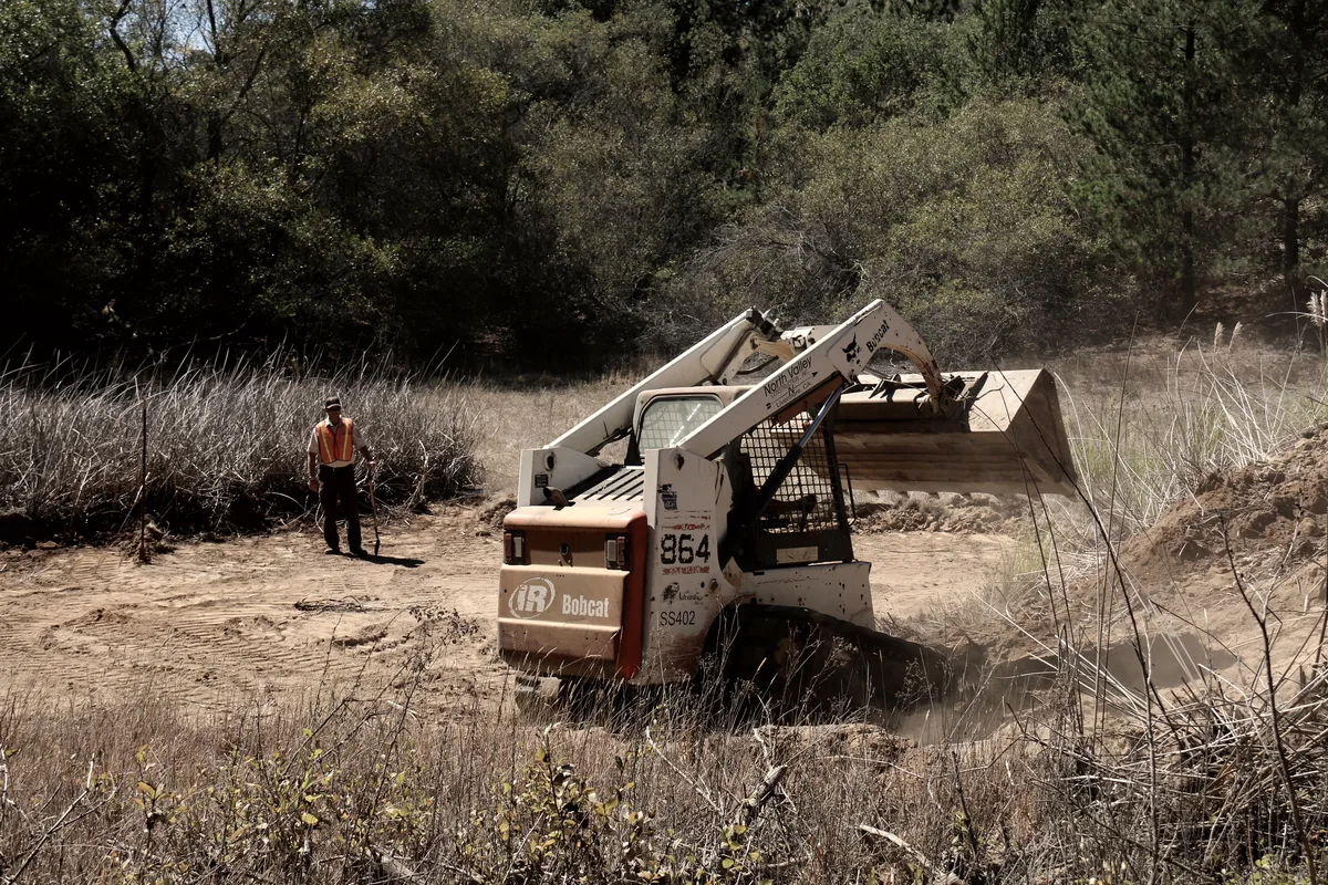 A skid-steer excavating the basin of a new pond in a brushy field, a worker standing by.